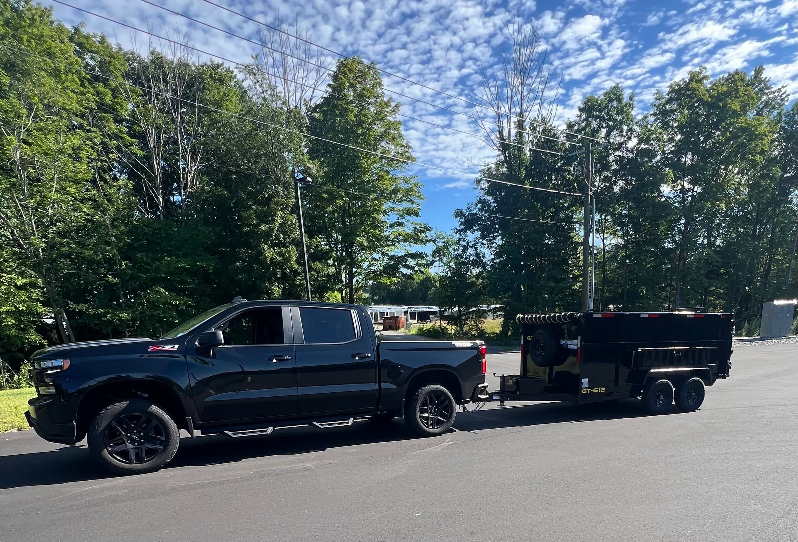 Black Chevrolet Silverado Z71 towing a black dump trailer on an asphalt road with trees.