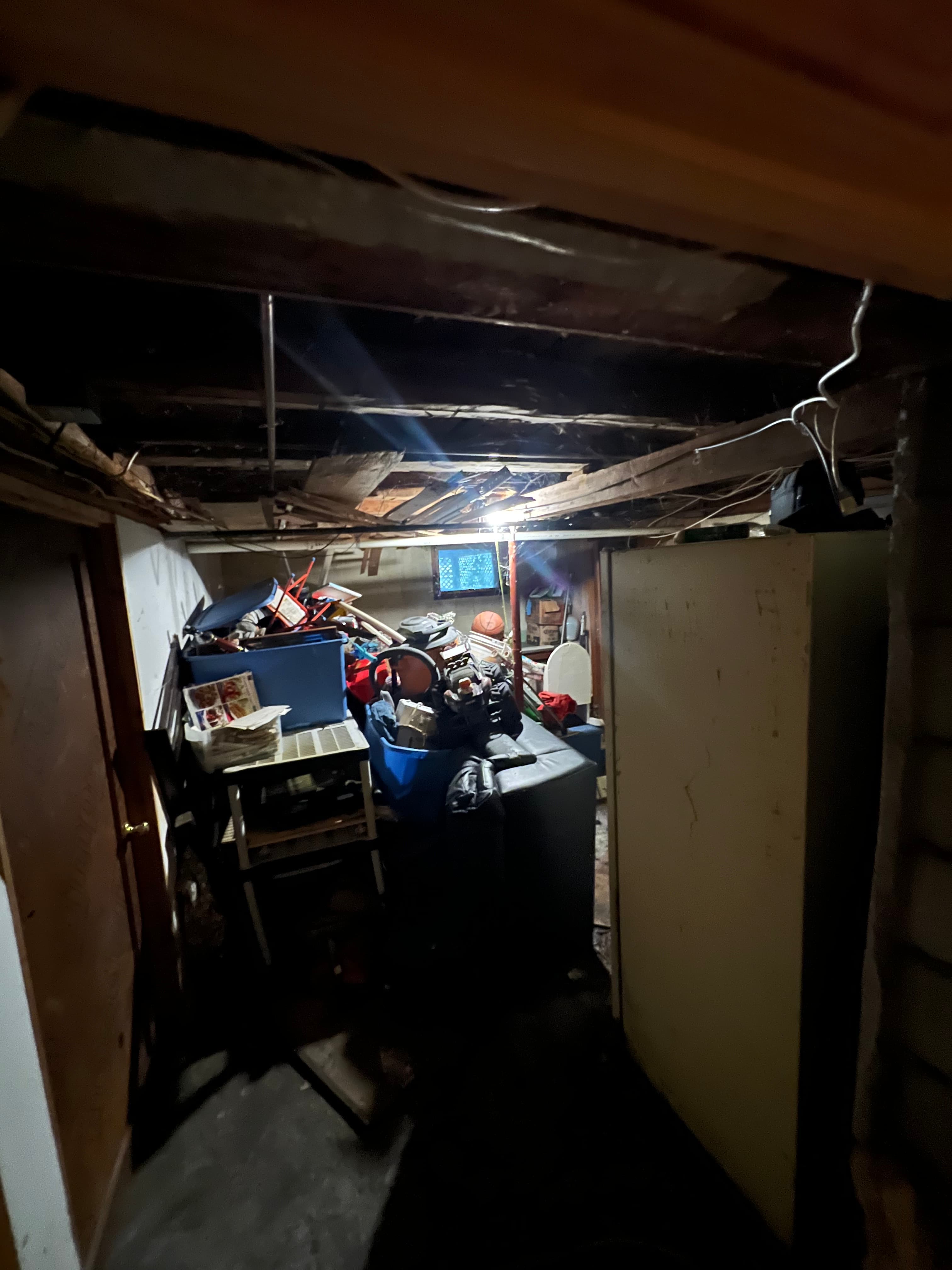 Dark, cramped basement filled with overflowing storage bins, old furniture, and exposed wooden ceiling joists.
