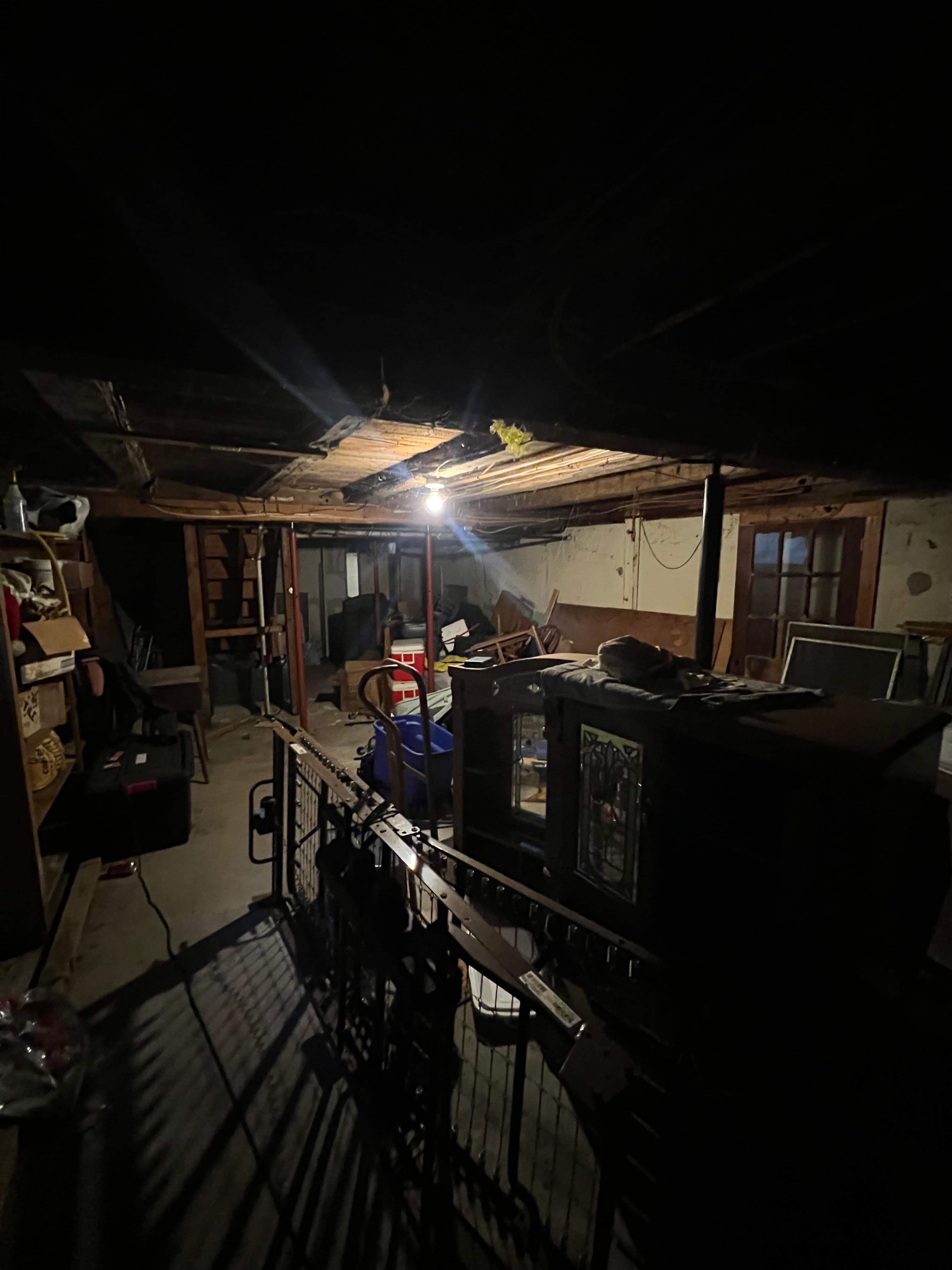 Dark, cluttered basement with exposed beams and furniture lit by a single hanging bulb.