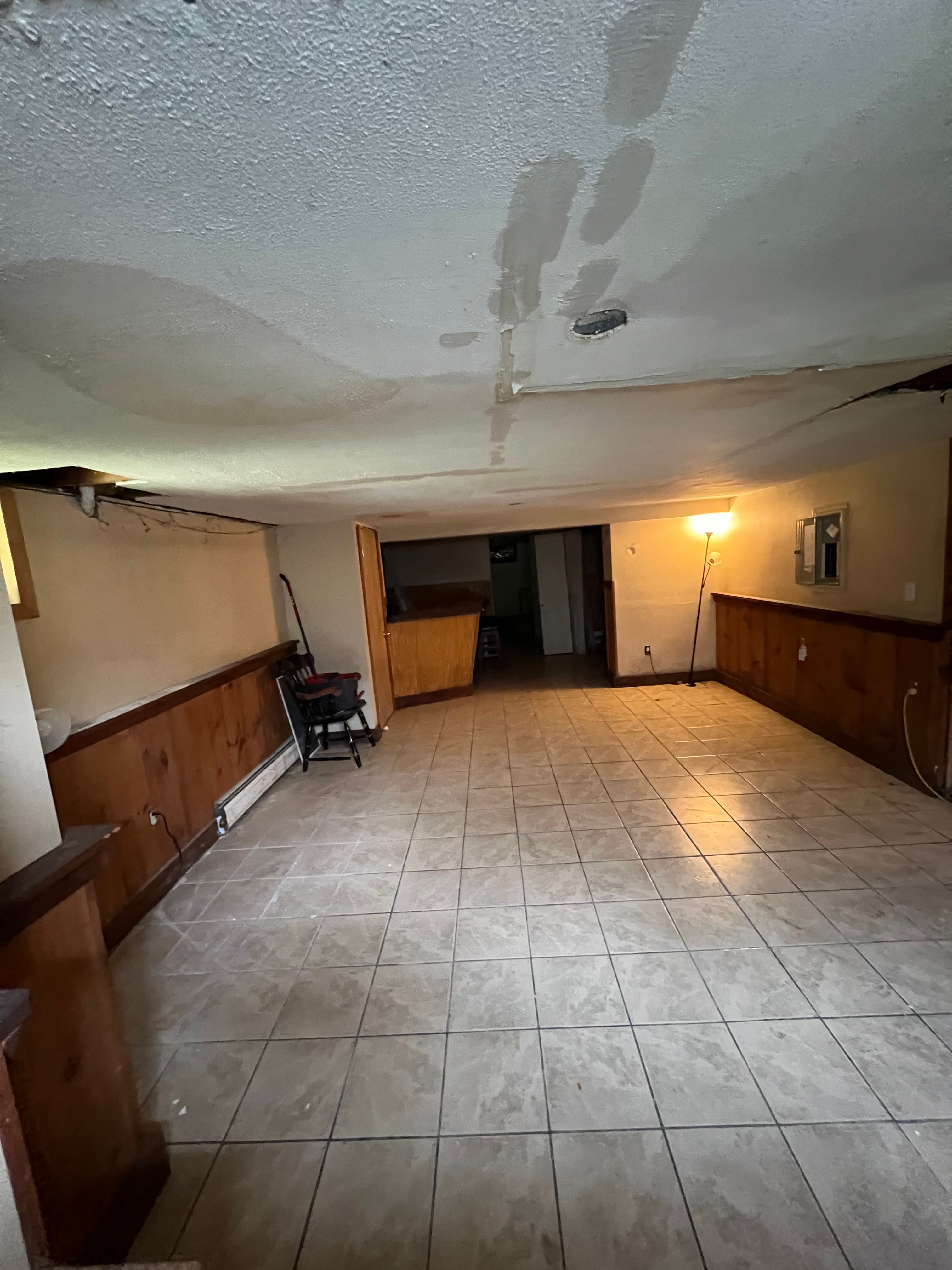 Empty basement room with tiled floors, wood-paneled walls, and a ceiling showing significant water damage.