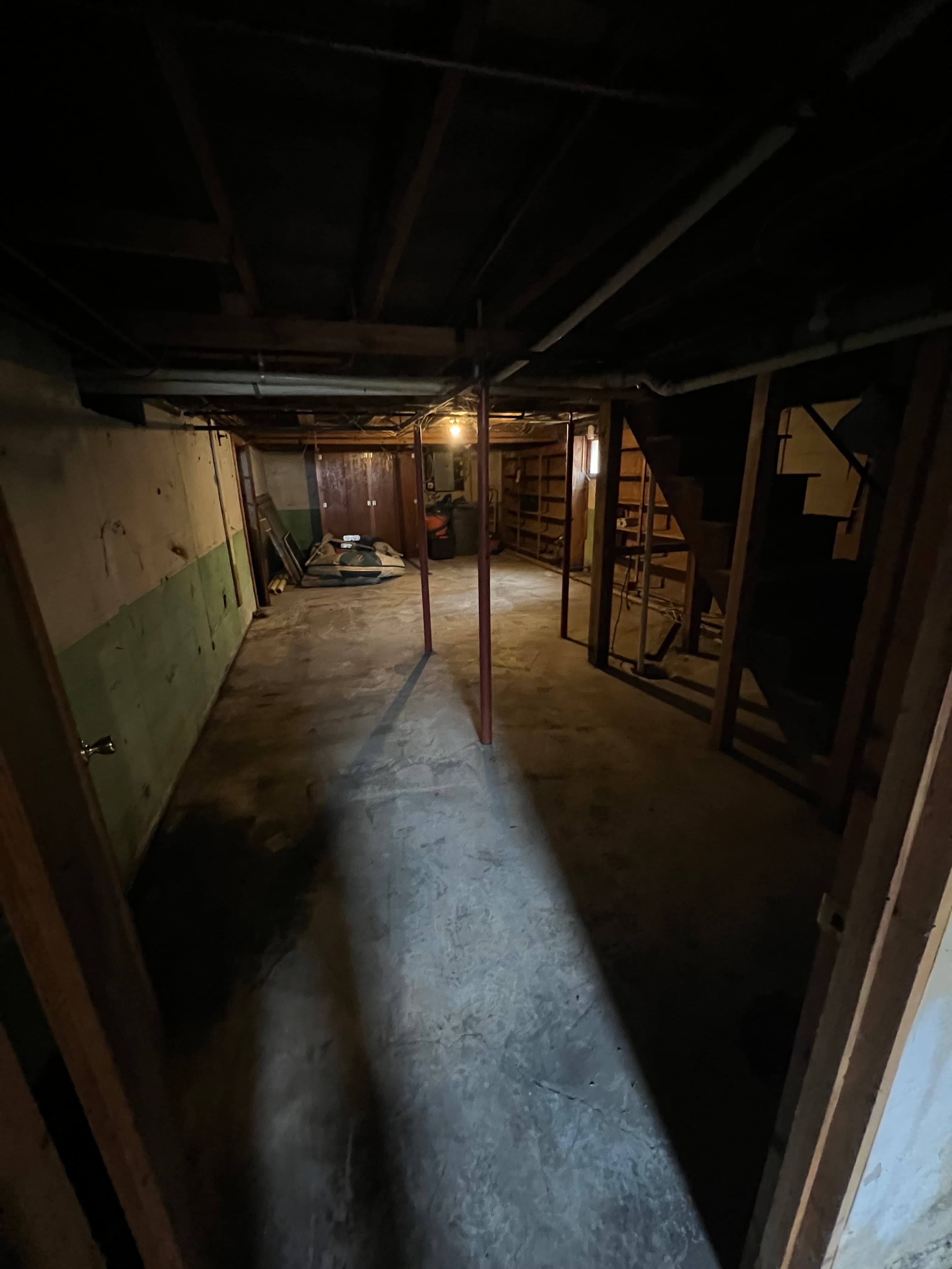 Dimly lit unfinished basement with concrete floors, wooden ceiling joists, and metal support poles.